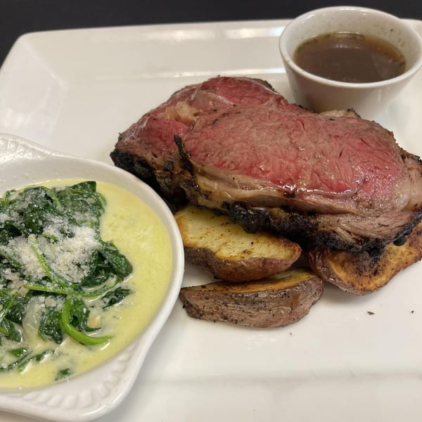 Prime rib with roasted potatoes, creamed spinach, and a side of au jus on a white plate.