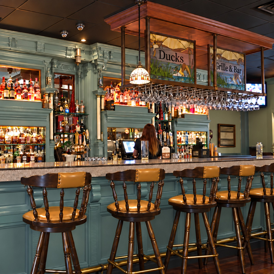 Elegant restaurant bar with teal paneling, liquor shelves, hanging wine glasses, duck-themed stained glass, and wooden bar stools.