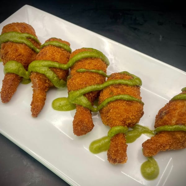 Five golden-brown fried chicken lollipops drizzled with green sauce on a white rectangular plate.