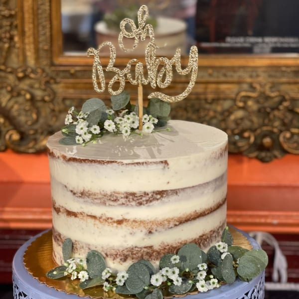 Naked cake with white frosting, eucalyptus, small white flowers, and a glittery "Oh Baby" topper.