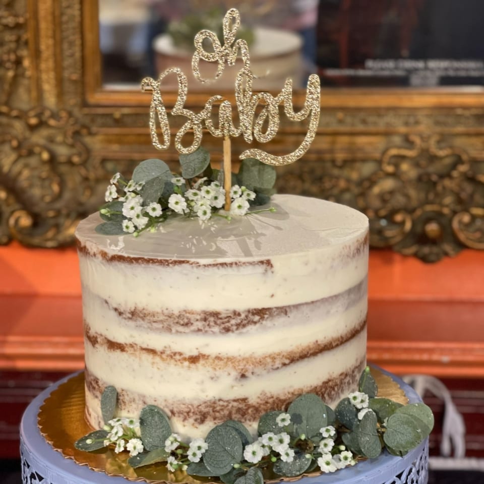 Naked cake with white frosting, eucalyptus, small white flowers, and a glittery "Oh Baby" topper.