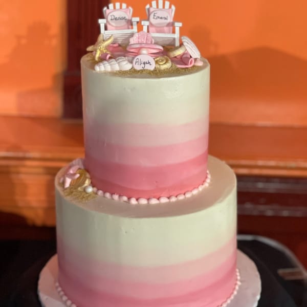 Two-tier pink and white ombre cake with beach-themed decorations and names "Denise," "Emerald," and "Aliyah."